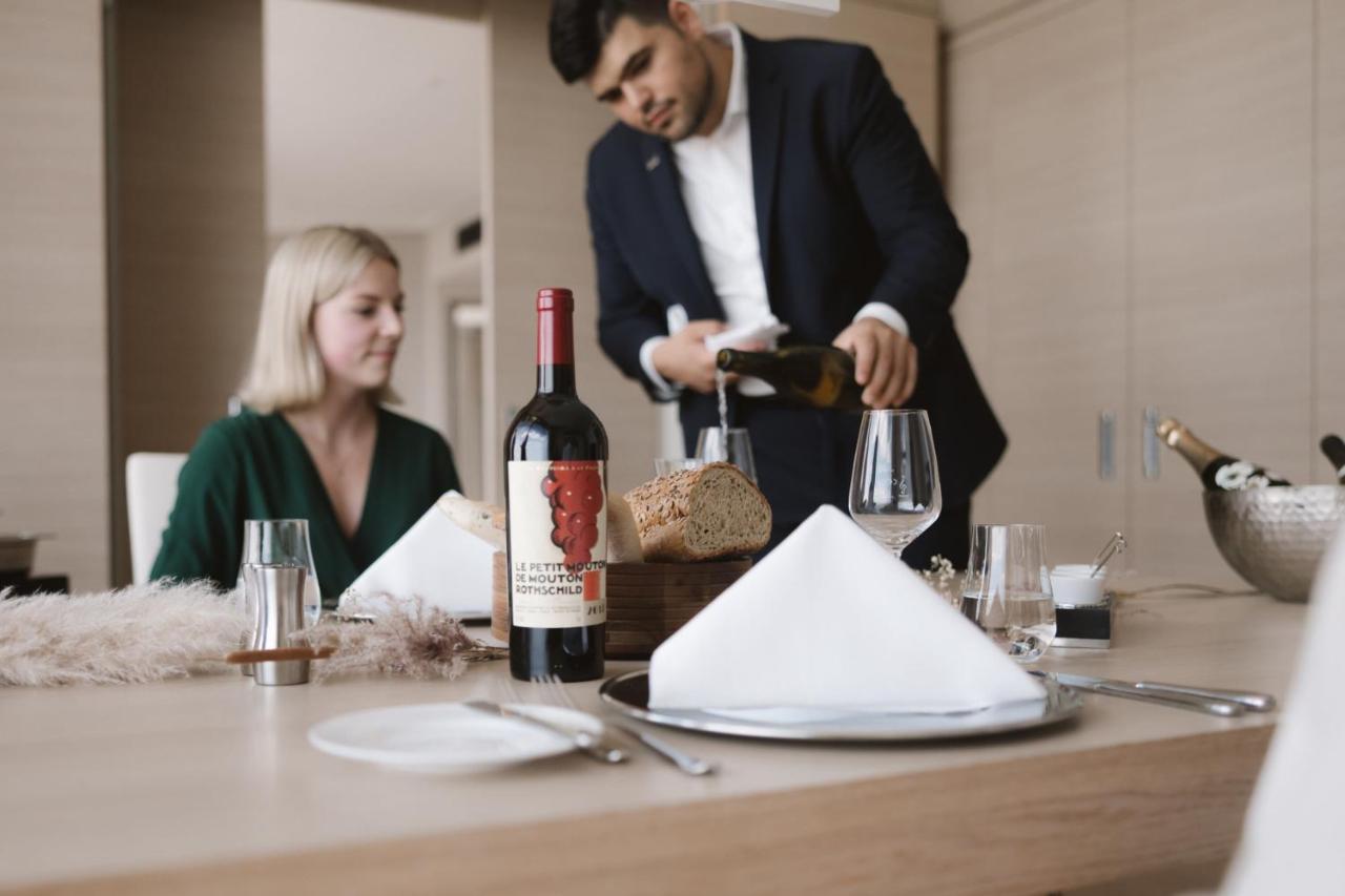 The Westin Hamburg Elbphilharmonie Hotel Luaran gambar The photo shows a dining scene featuring a well-dressed man pouring wine into a glass while a woman sits at the table. The table is elegantly set with a folded napkin, wine glasses, and a bottle of red wine placed prominently. There is also a loaf of