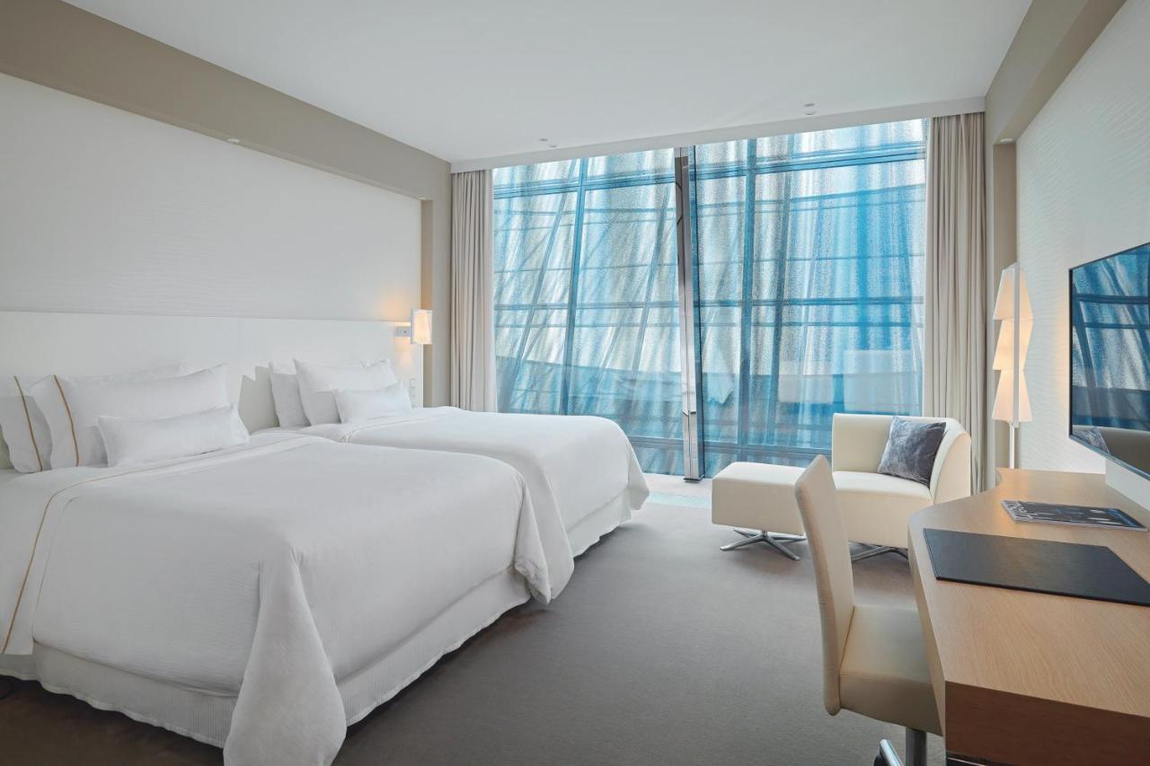 The Westin Hamburg Elbphilharmonie Hotel Luaran gambar The photo shows a modern hotel room featuring two white beds with plush bedding. There is a large window with sheer curtains that allows natural light to filter in, offering a view outside. The room has neutral-colored walls and carpeting, creating a