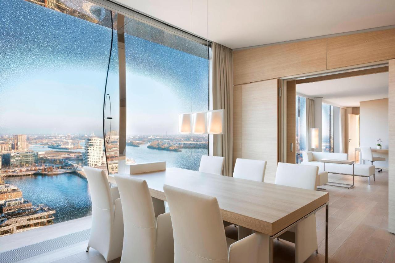 The Westin Hamburg Elbphilharmonie Hotel Luaran gambar The photo shows a modern dining area with a large, light-colored table surrounded by white chairs. The space features a panoramic window that offers a stunning view of a body of water and a cityscape. The decor is minimalistic and elegant, with smoot