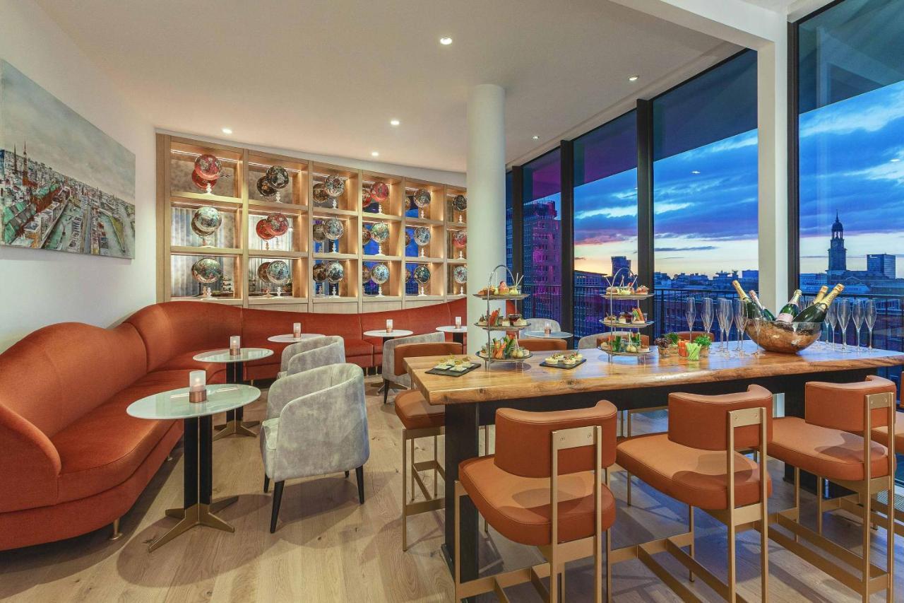 The Westin Hamburg Elbphilharmonie Hotel Luaran gambar The photo shows a modern and stylish restaurant or café interior. It features a combination of comfortable seating, including a curved, orange sofa and gray chairs. The tables are round with light-colored tops, and there are several tiered stands dis