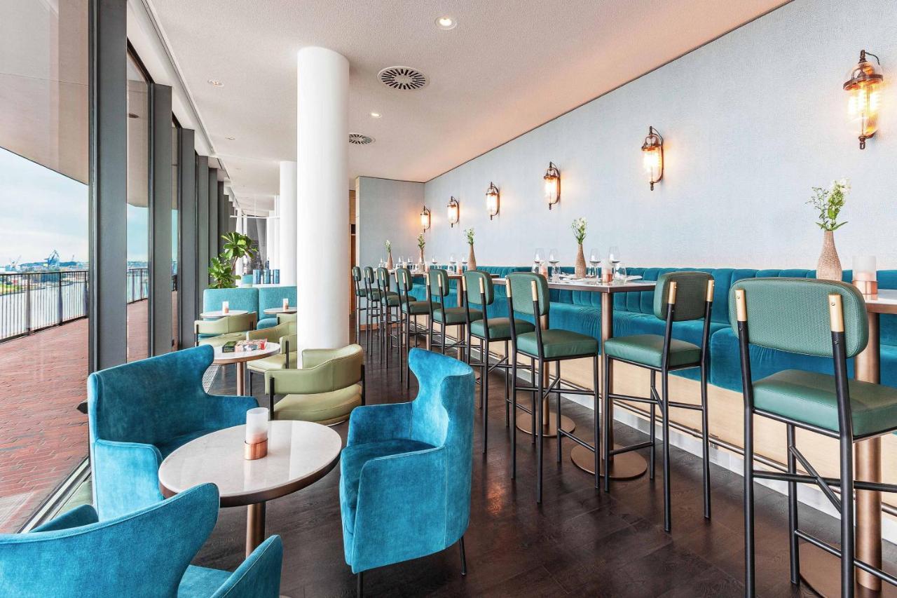 The Westin Hamburg Elbphilharmonie Hotel Luaran gambar The photo shows a modern and stylish restaurant or café interior. It features a long row of high-backed bar stools at a counter, complemented by plush seating in the form of rounded chairs and small tables arranged throughout the space. The color sch