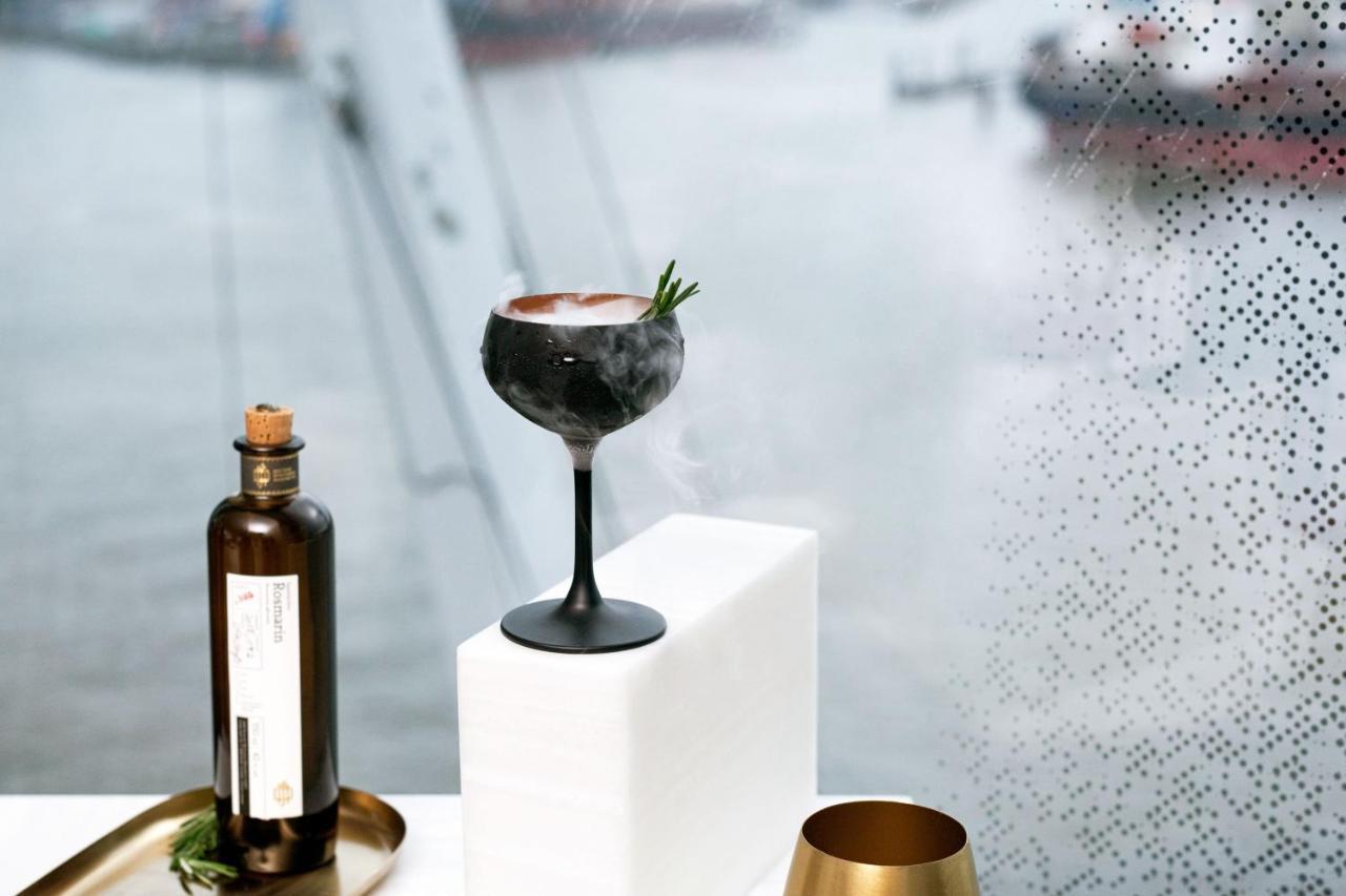 The Westin Hamburg Elbphilharmonie Hotel Luaran gambar The photo features a stylish cocktail elegantly presented on a white pedestal. The cocktail is served in a sophisticated black stem glass, topped with a garnish of rosemary. Beside it, there is a dark glass bottle, likely containing a spirit or mixer