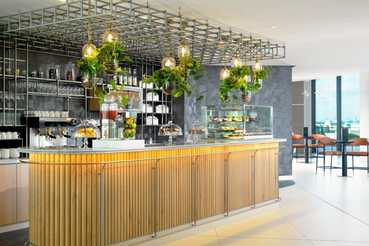 The Westin Hamburg Elbphilharmonie Hotel Luaran gambar The photo shows a modern café or bar setting with a sleek wooden counter that features vertical slats. Above the counter, there is a grid-like framework hanging from the ceiling, adorned with various green plants that add a touch of nature to the int