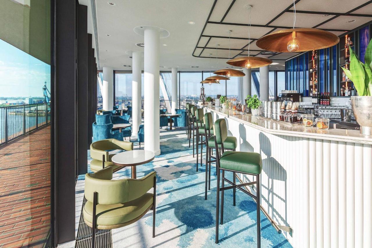 The Westin Hamburg Elbphilharmonie Hotel Luaran gambar The photo depicts a modern bar or lounge area with a stylish design. There are high-backed green chairs surrounding small circular tables, arranged in an open space that has large windows showcasing a view of the outside. The bar area is sleek, featu