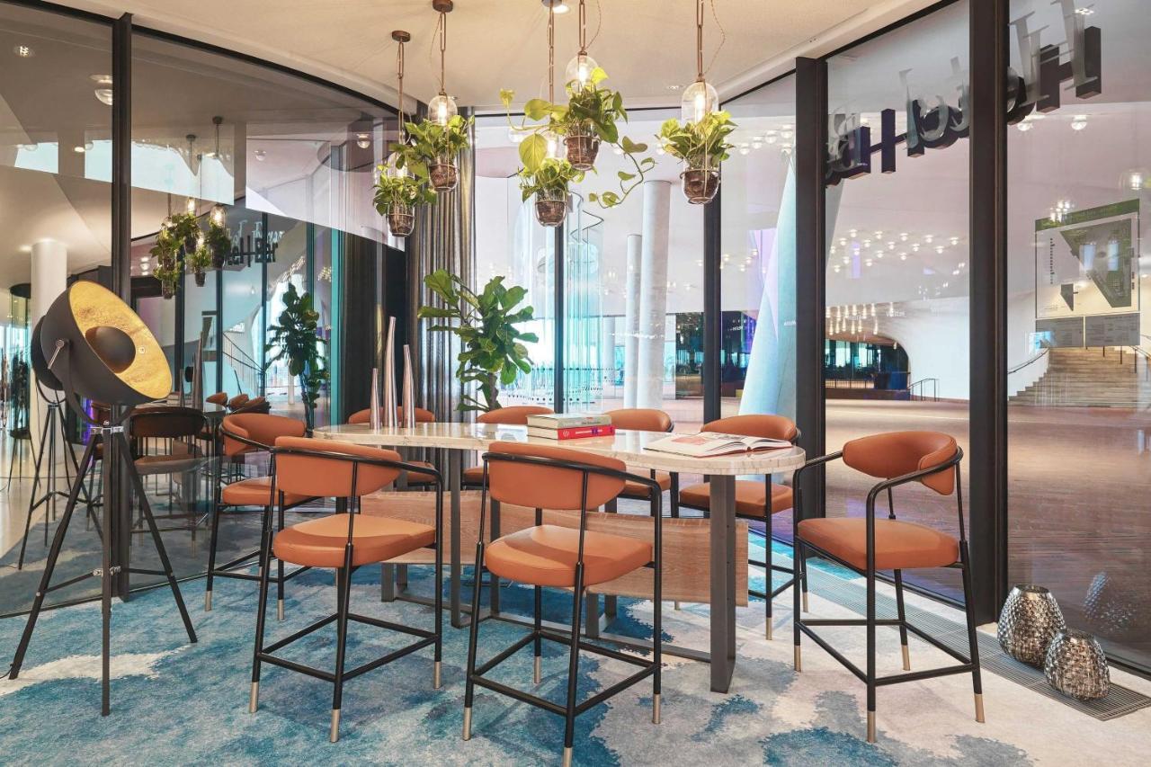 The Westin Hamburg Elbphilharmonie Hotel Luaran gambar The image depicts a modern indoor seating area designed for gatherings or meetings. There is a long table surrounded by several bar-style chairs with brown upholstery. Above the table, there are hanging plants that add a touch of greenery to the spac