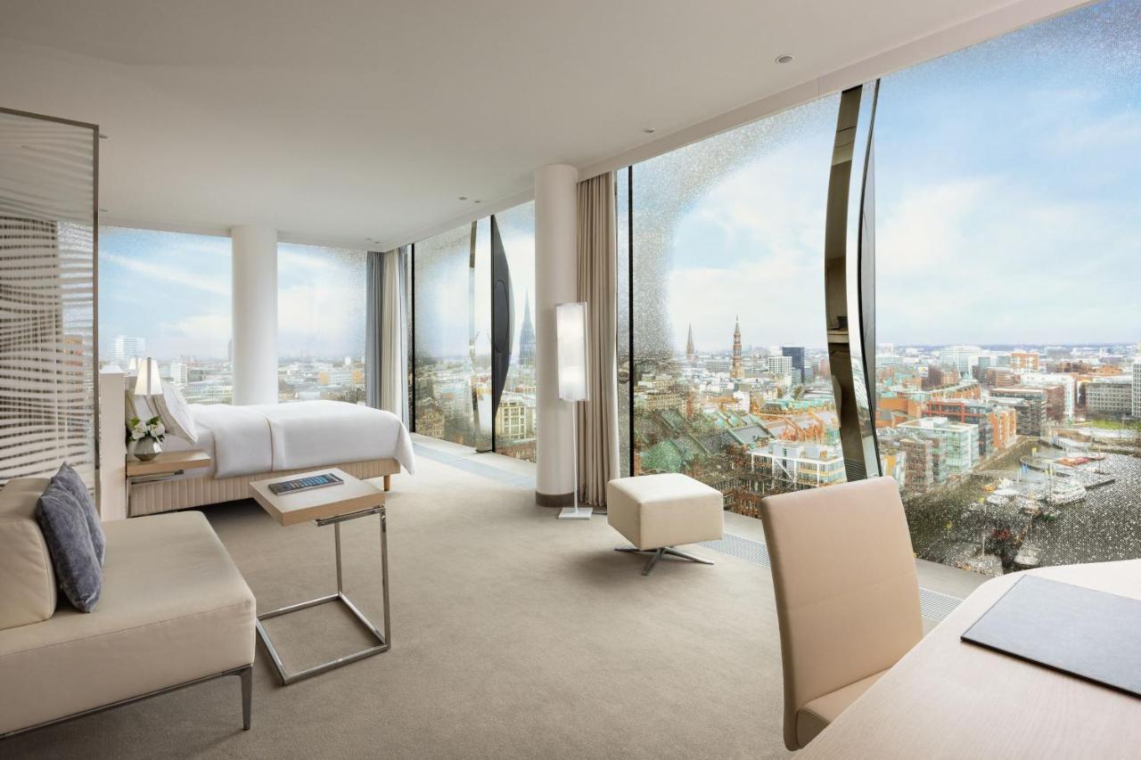 The Westin Hamburg Elbphilharmonie Hotel Luaran gambar The photo shows a modern hotel room with large floor-to-ceiling windows that provide a panoramic view of a city landscape. The room features a minimalist design with neutral tones. There's a king-sized bed positioned in the center, a sleek desk and c