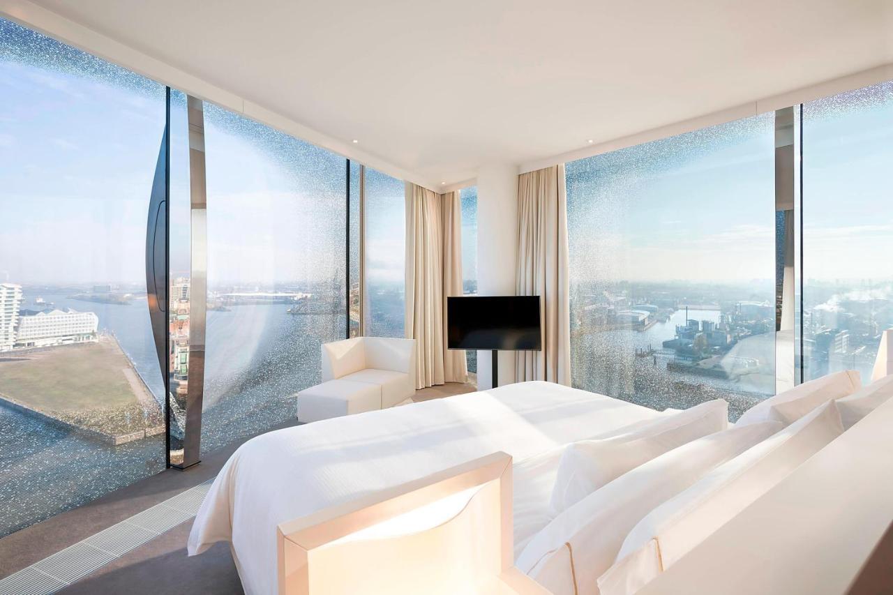 The Westin Hamburg Elbphilharmonie Hotel Luaran gambar The photo shows a modern hotel room featuring a large bed with white linens and a minimalist design. It has expansive floor-to-ceiling windows that offer a panoramic view of the landscape outside, likely a city or waterfront setting. The room is deco