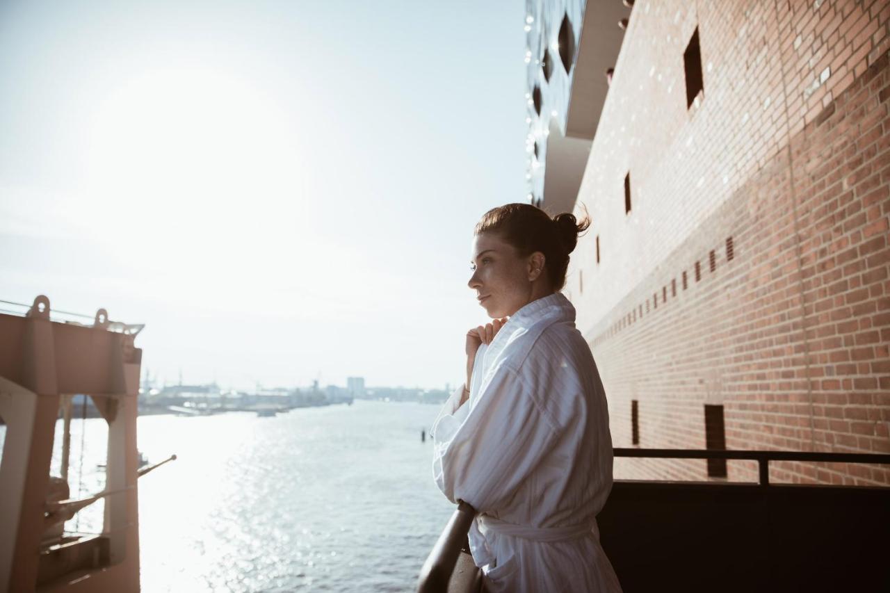 The Westin Hamburg Elbphilharmonie Hotel Luaran gambar The photo depicts a person standing on a balcony, looking out over a body of water. The individual is wearing a white bathrobe, and their hair is styled in a bun. The background features a scenic view with sunlight illuminating the scene, creating a 