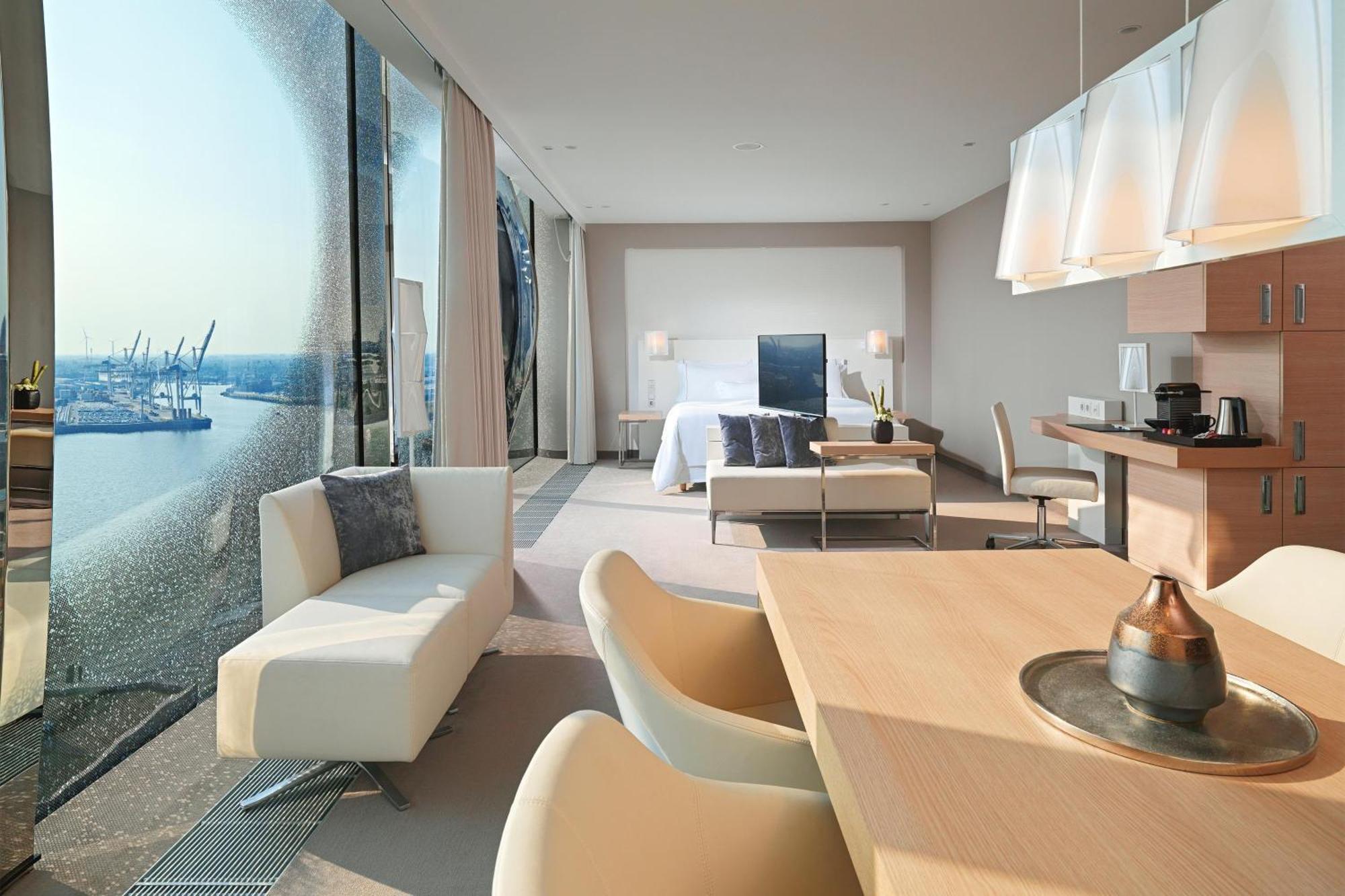 The Westin Hamburg Elbphilharmonie Hotel Luaran gambar The photo shows a modern hotel room with a spacious layout. On one side, there are large floor-to-ceiling windows that offer a view of the outside, likely showcasing a waterfront or cityscape. Inside, there's a bed with a minimalist design, accompani