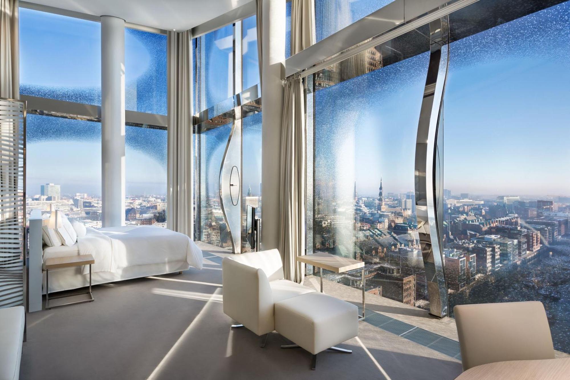 The Westin Hamburg Elbphilharmonie Hotel Luaran gambar The photo shows a modern hotel room with a stunning, panoramic view through large, uniquely designed windows. The room features a sleek, minimalist aesthetic with light-colored furnishings, including a white bed and a couple of chairs. The walls are 